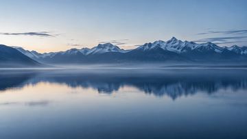 Lac calme au crépuscule, devant une chaîne de montagnes enneigées.